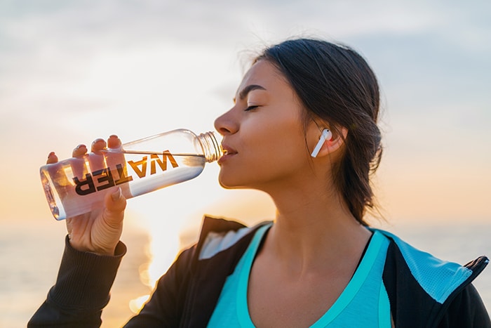 attractive slim woman doing sport exercises on morning sunrise beach in sports wear, thirsty drinking water in bottle, healthy lifestyle, listening to music on wireless earphones, smiling happy